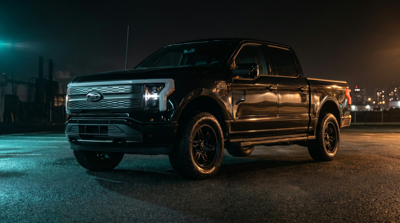 Modern black pickup truck photographed at night with dramatic teal and amber rim lighting on an empty lot